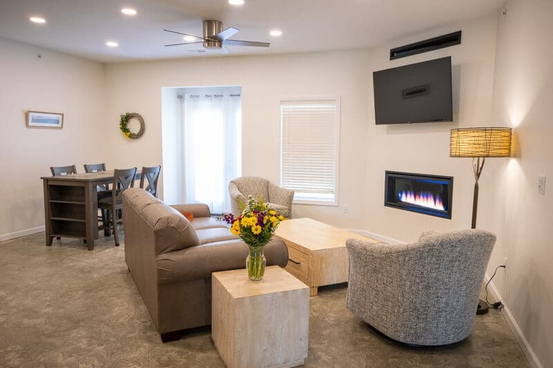 A cozy living room featuring a sofa, two chairs, a dining table, a modern fireplace, and a vase of flowers on a wooden coffee table.
