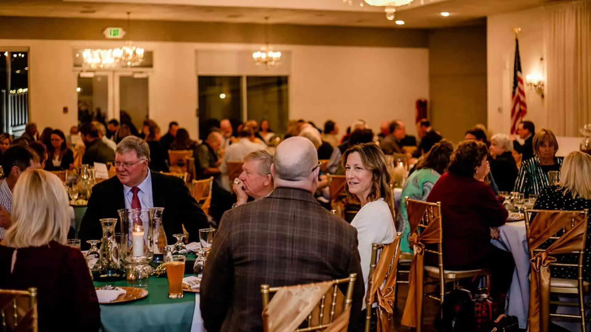 Large banquet hall with gold chairs, tables with green tablecloths, full place settings, and people eating and talking with each other