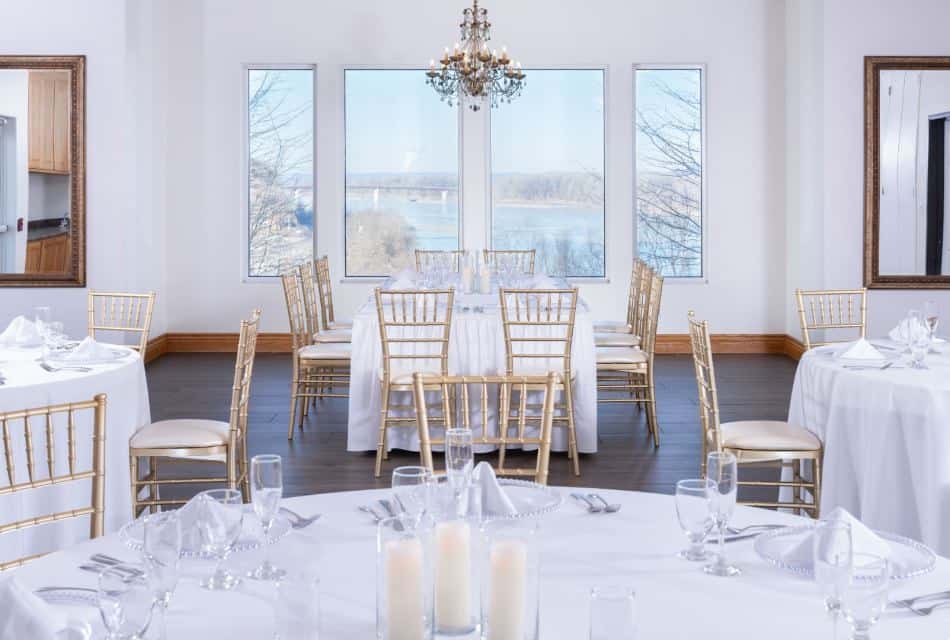 Large banquet hall with gold chairs, tables with white tablecloths, full placesettings, and windows showing the river