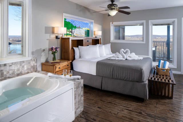 A cozy hotel room featuring a large bed, a spa tub, and large windows with scenic views.