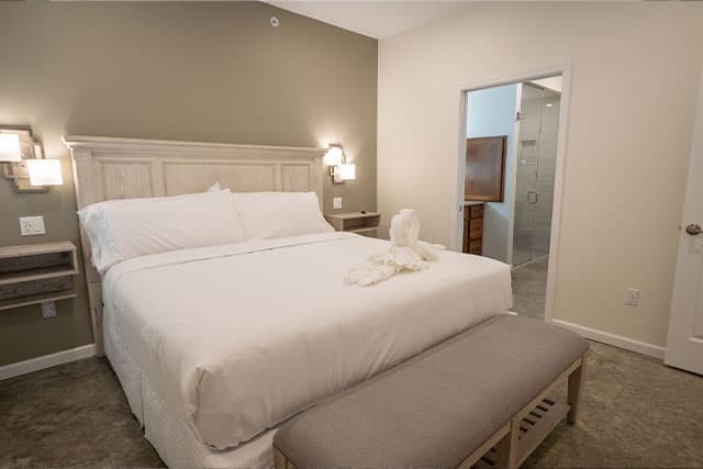 A modern bedroom with a large bed, a bench at the foot, and an adjacent bathroom entrance.