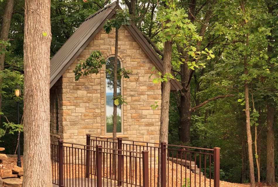 Small stone building with arched window in the woods and lots of trees in background. Metal railed footbridge and tree trunks in foreground.