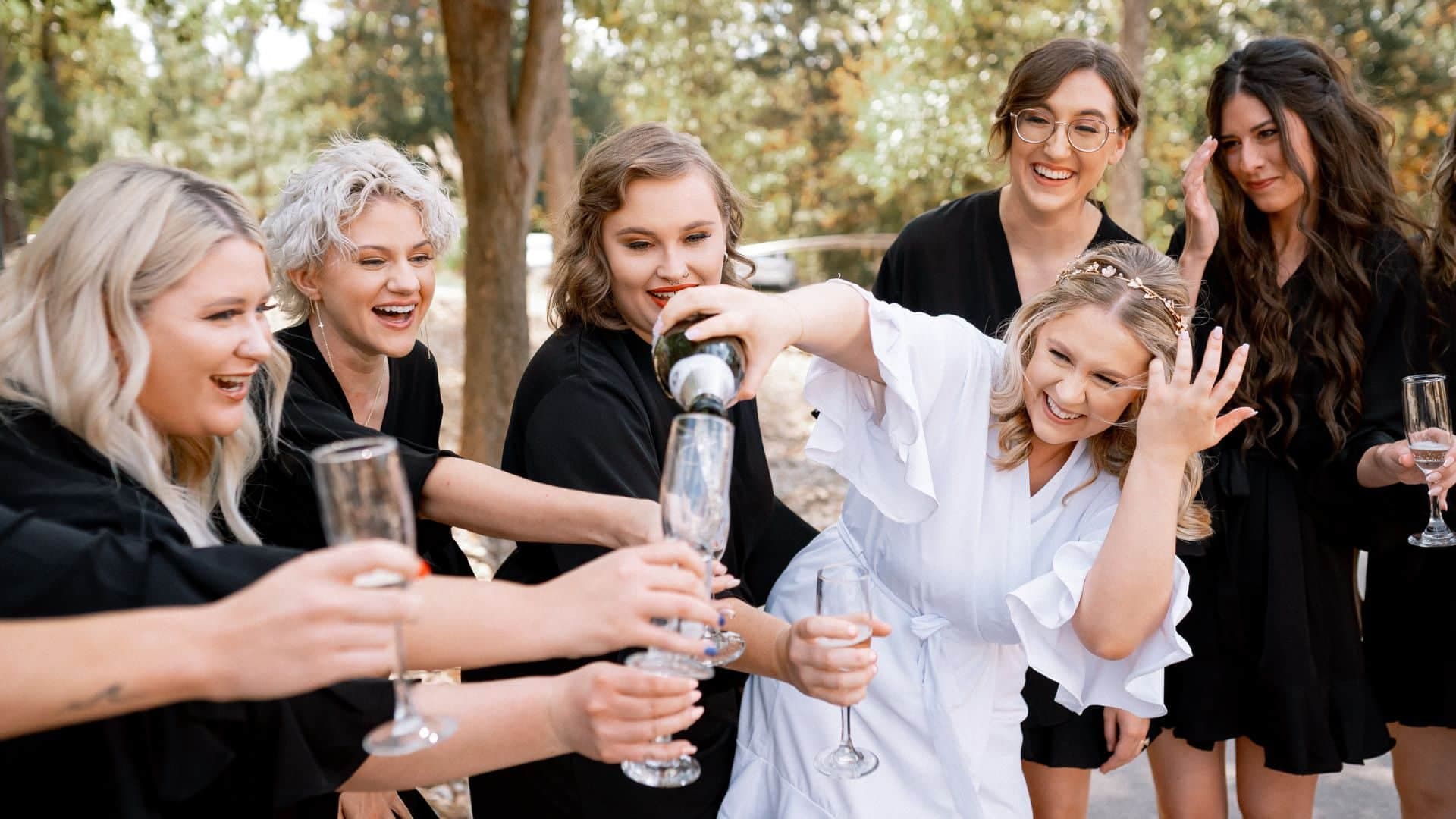 A group of girls wearing black bathrobes smiling at each other and holding flutes while bride dressed in a white robe pours champagne