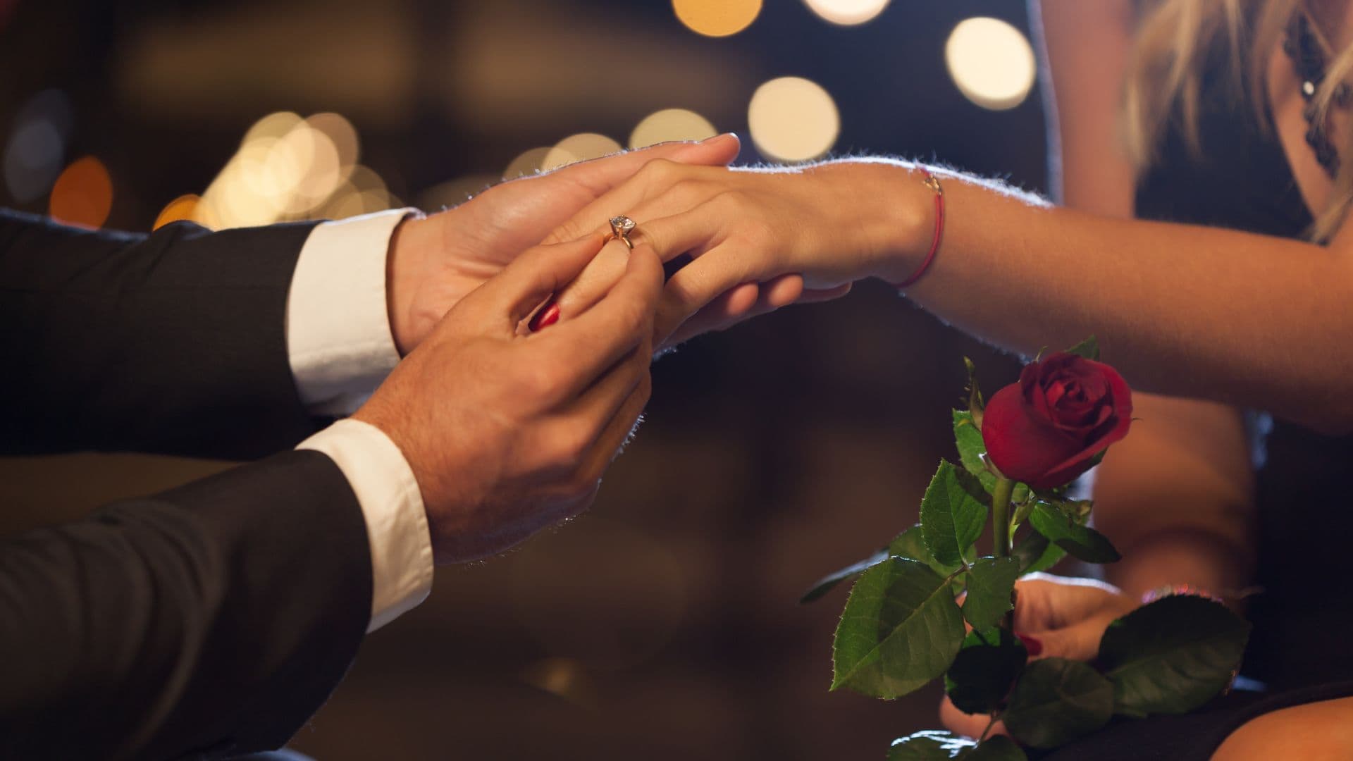 Man in suit putting a diamond ring a lady's finger with twinkle lights in the background