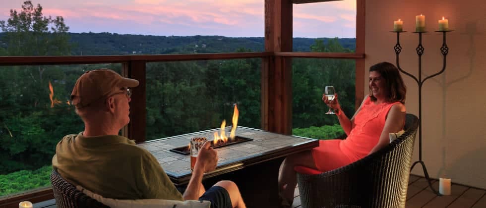 a man and woman relax by a fire-pit at dusk while drinking wine