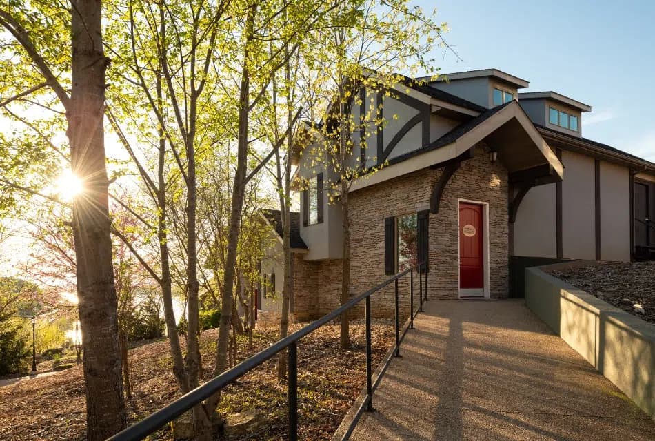Exterior view of one of the cottages with pathway leading up to red door surrounded by trees and sun shining in the background
