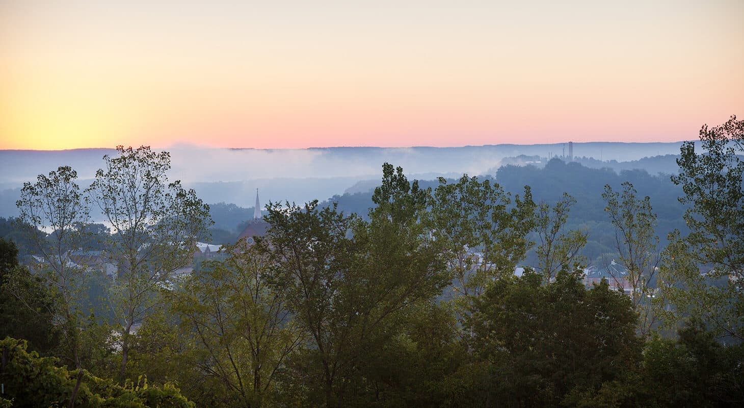 A serene landscape at sunrise with mist rolling over hills and trees in the foreground.