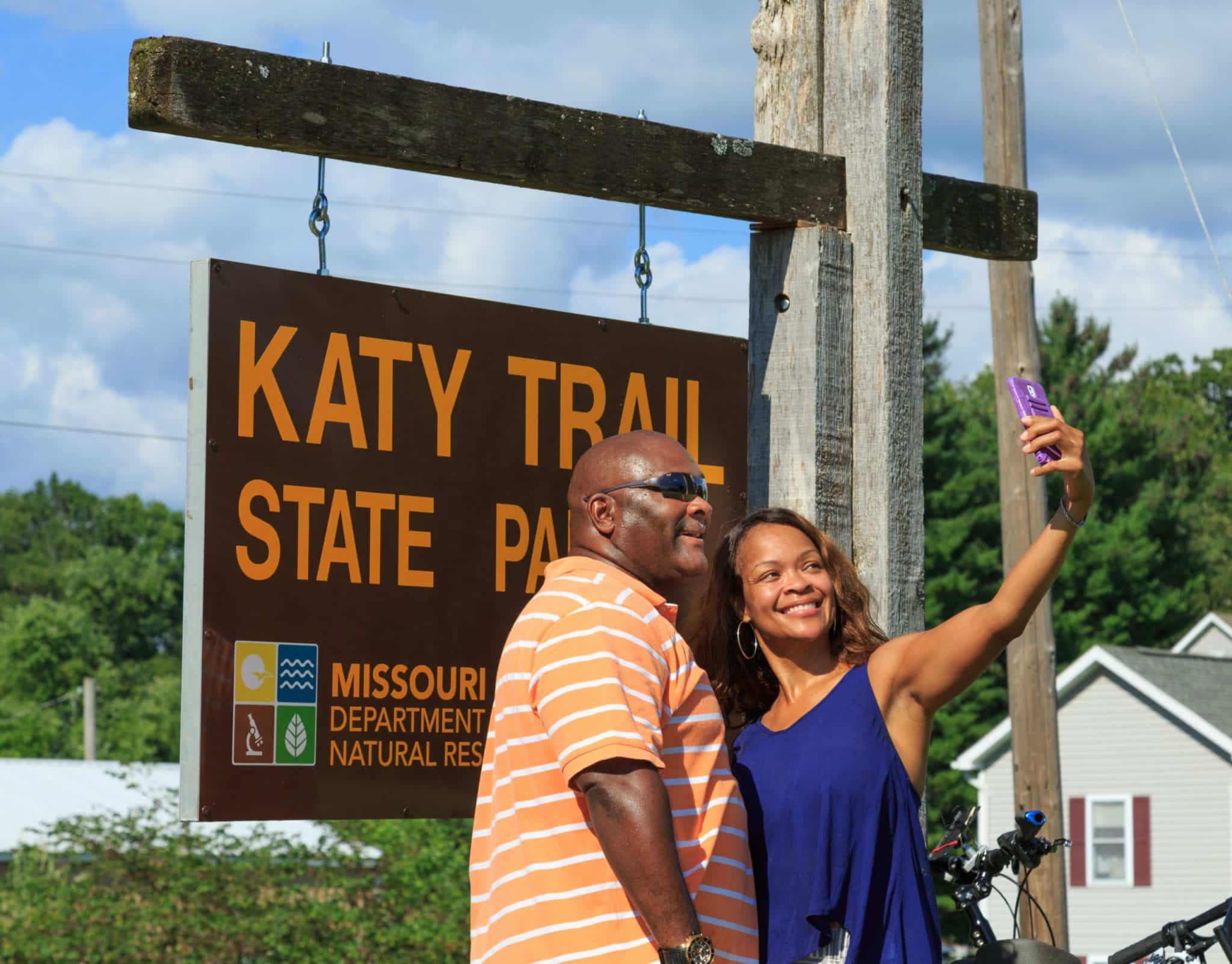 A man and a woman pose for a selfie in front of the Katy Trail State Park sign.