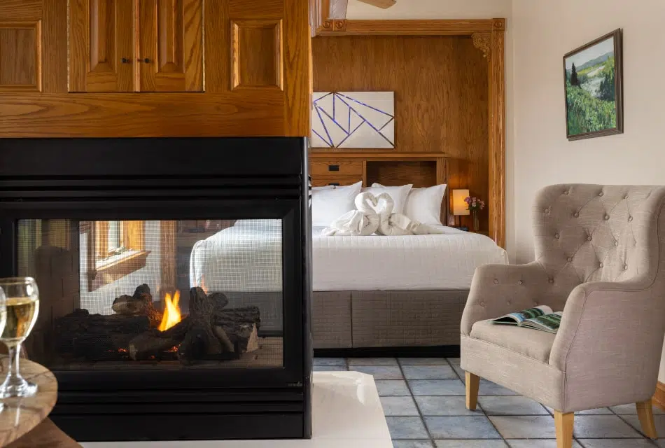 Large bedroom suite with cream walls, tiled flooring, see through fireplace, wooden headboard, white bedding, and light taupe upholstered chair