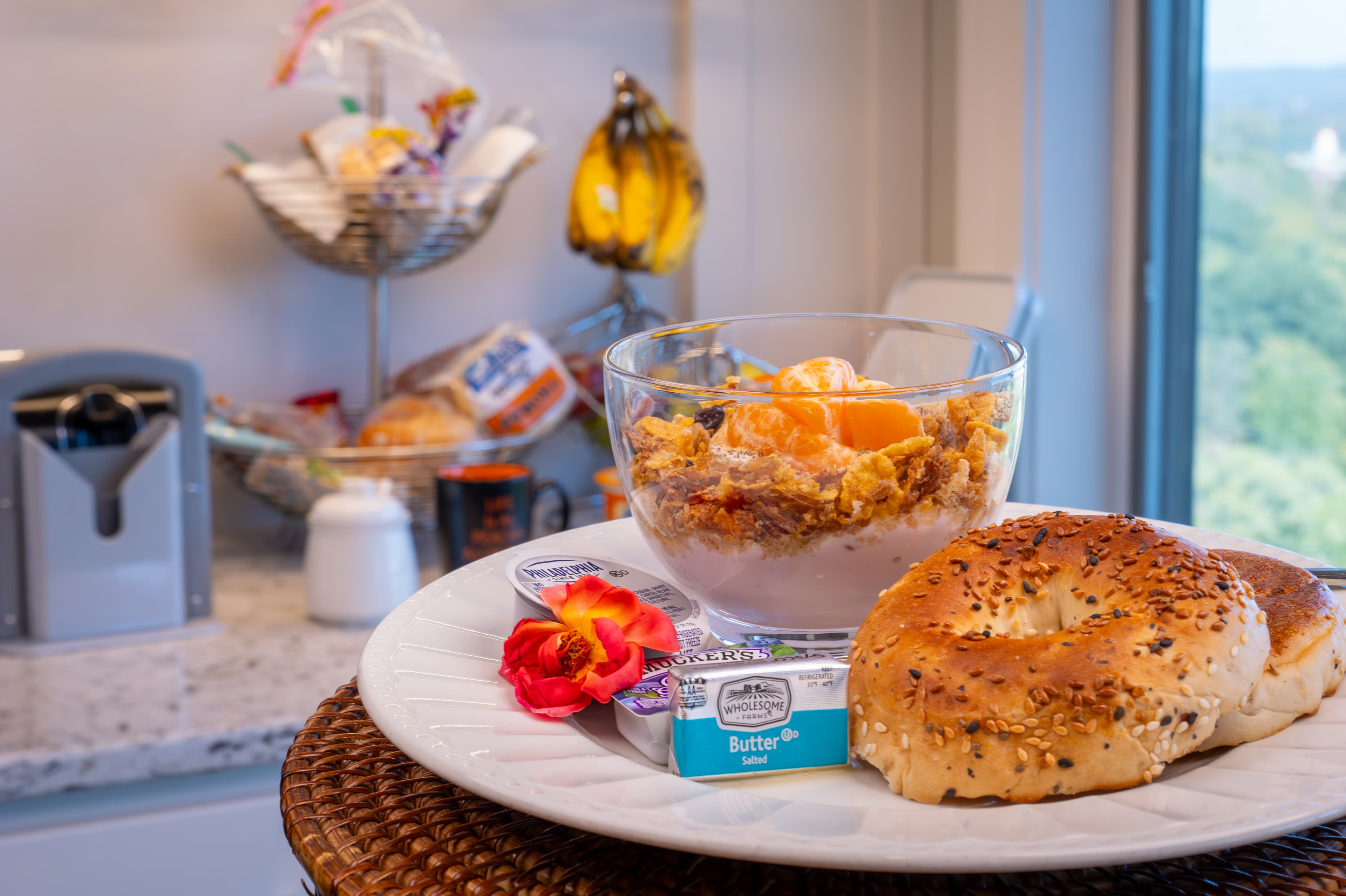Breakfast plate in foreground of picture. Plate has a bagel sliced in half with butter and cream cheese packets on plate. There is also a bowl of cereal with fruit in a clear bowl.