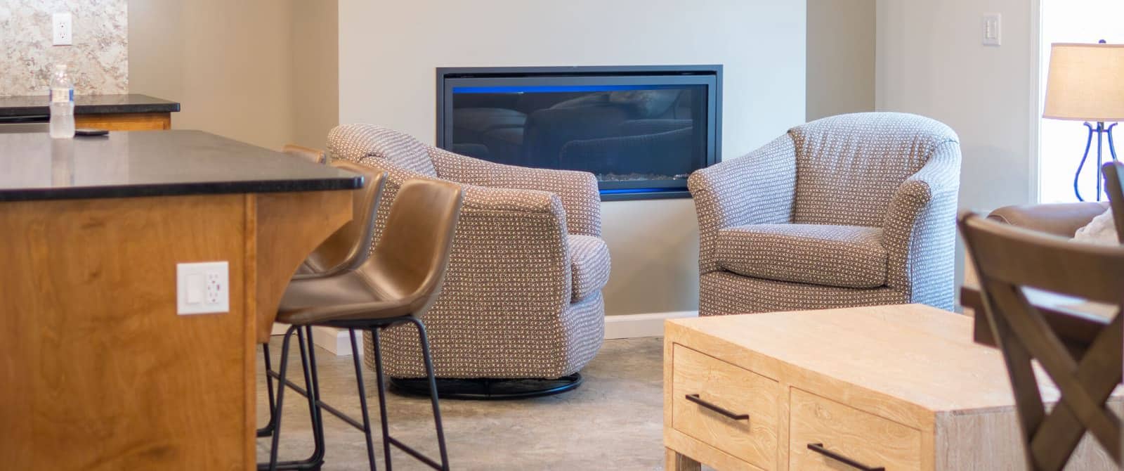 View of living room with inset fireplace and kitchen with island and brown cushioned chairs.