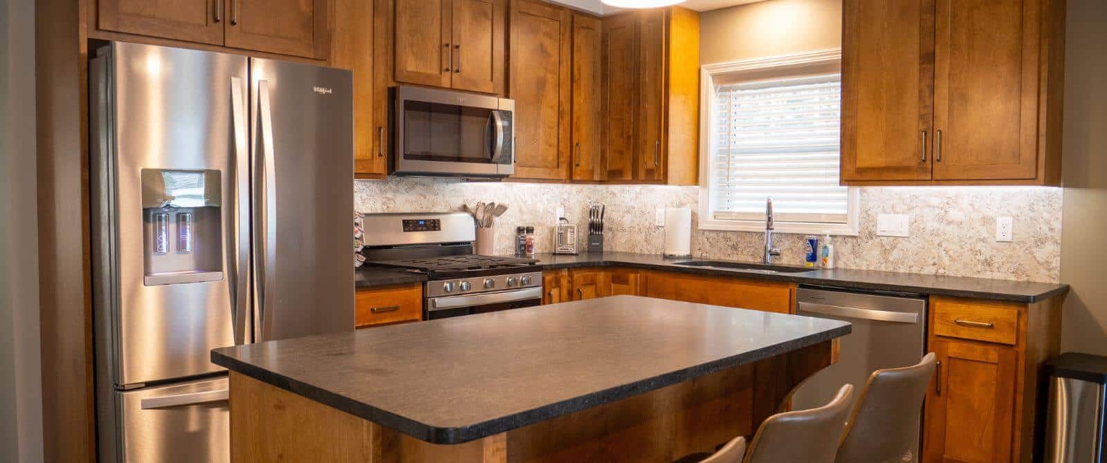 Closeup view of kitchen with island with cushioned chairs, stainless steel appliances, a lot of wooden cabinets and window