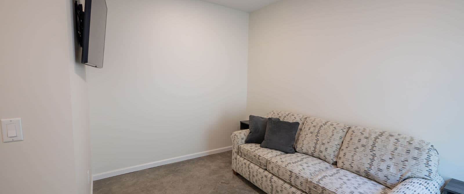 narrow room with cream couch with brown carpet and flat tv hanging on the wall