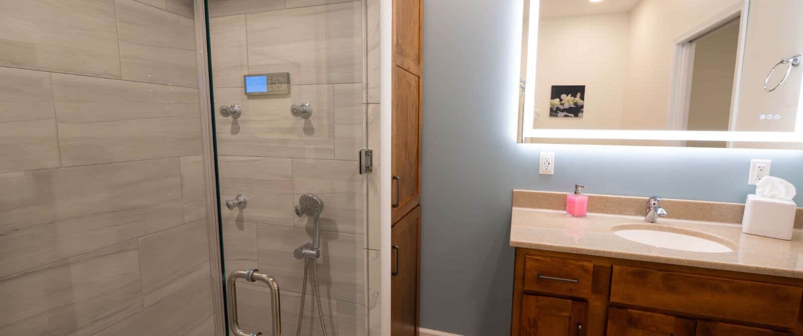 Beige colored tiled glass enclosed shower with beige vanity with wood cabinets with mirror hanging above.