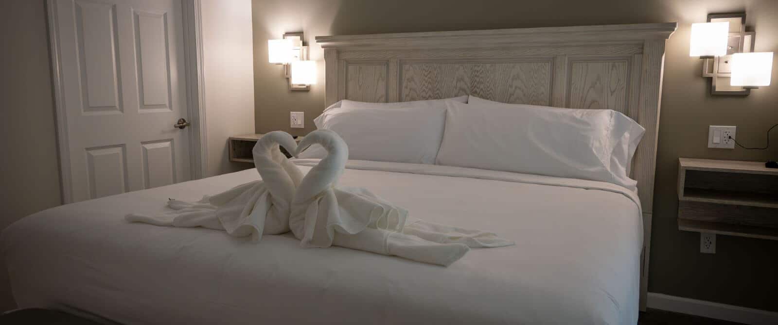 King bed with white coverings with towels in form of swans on it, nightstands with lights hanging above