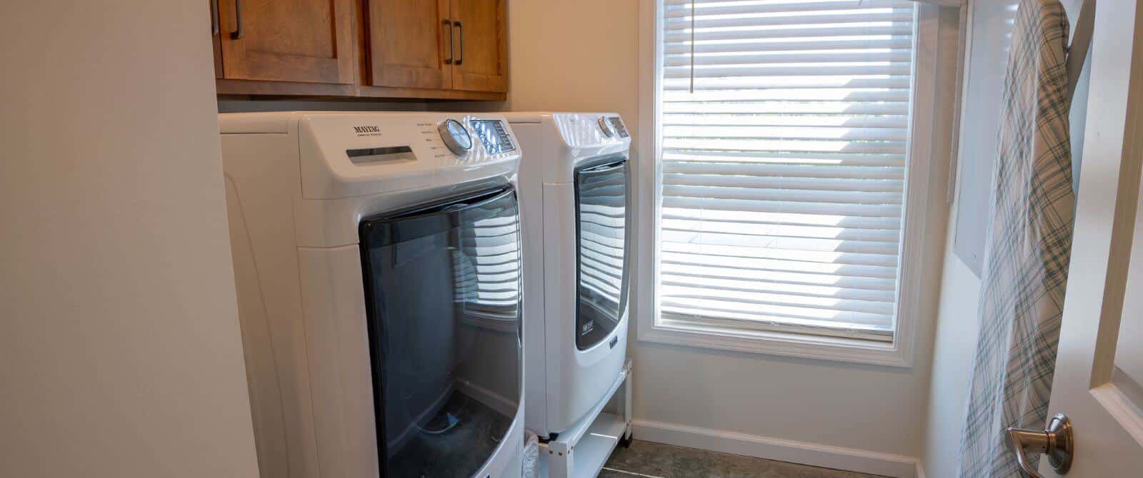 Laundry room with side-by-side white appliances with cabinets above, window with mini blinds, ironing board hanging on wall