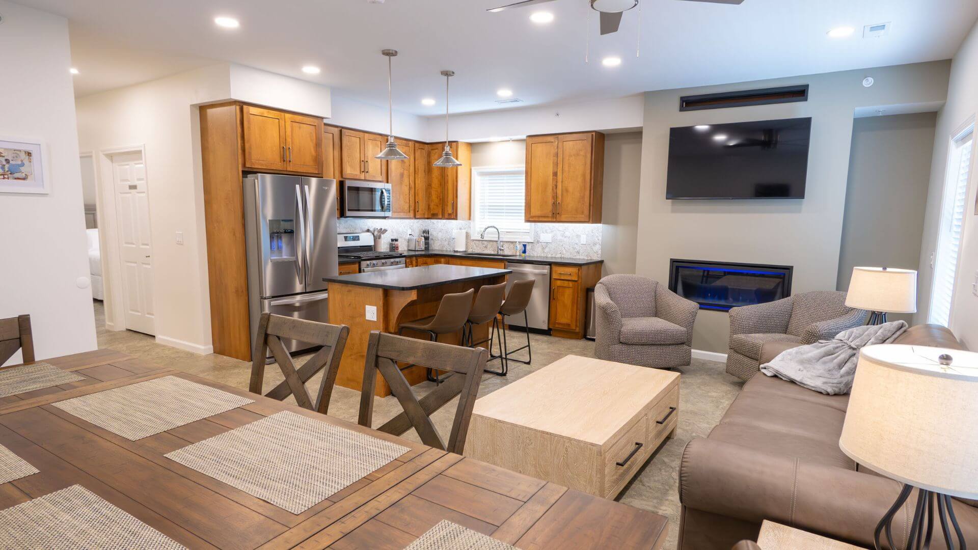 View of 3 rooms - kitchen with stainless appliances and a lot of cabinets, dining room table and living room with couch & chairs with Tv hanging on wall above inset fireplace.