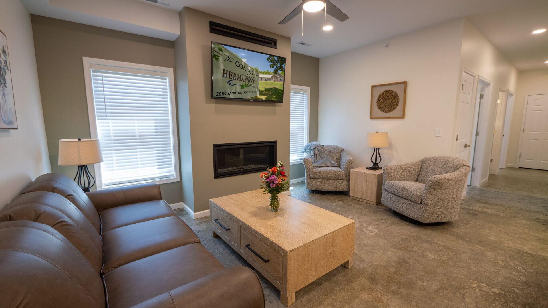 Large living room with windows, brown leather couch and other chairs with flat screened tv on wall above inset fireplace